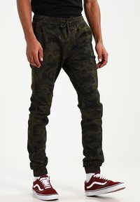 Maskovací joggingové kalhoty v tmavě zelené barvě se elastickým pasem a náplety na kotnících. Vzhledem k červeným a bílým teniskám.