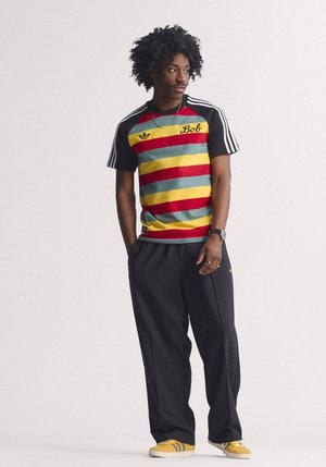 Homme aux cheveux bouclés portant un t-shirt Adidas rayé avec l'inscription "Bob", un pantalon à fines rayures, des baskets Adidas jaunes, une montre et une chaîne, debout les mains dans les poches.