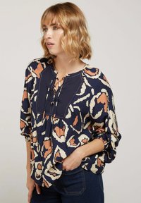 Blouse navy avec imprimé animal en crème et rouille. Présente un col rond avec des liens, des manches bouffantes et une coupe ample. Texture de tissu douce.