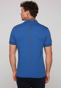 Camisa polo de algodón azul con un diseño texturizado, con detalles en negro en las mangas y la parte trasera, que muestra logotipos y texto impresos.