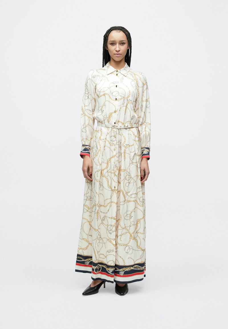 Gara balta krekla kleita ar zelta ķēdes rakstu, ar pogām priekšpusē, ar jostu viduklī un kontrastējošiem ziliem, sarkaniem un melniem akcentiem aprocēs un malā.