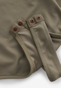 Material de culoare verdeoliv cu textură netedă; prezintă două curele cu butoane roșii circulare și cusături întărite de-a lungul marginilor.