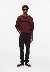 Maglione lavorato a maglia color marrone bordeaux con testo nero "KARL LAGERFELD", abbinato a jeans neri e sneakers nere, con lacci bianchi e suola in gomma.