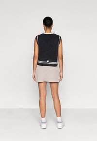 Bezrukávý tmavý pletený svetr s bílým horizontálním pruhem na lemu, kombinovaný s béžovým žebrovaným šaty a bílými teniskami. Hedvábně hladký povrch.