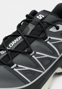 Pelēki skriešanas apavi ar teksturētu sieta augšdaļu, atstarojošiem akcentiem un strukturētu dizainu, melniem šņorēm un baltu logotipu.