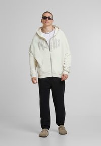 Beige Kapuzenjacke mit großem grauen Aufdruck, kombiniert mit schwarzen Jogginghosen und beigen Slippern. Sie hat einen lässigen Schnitt und Fronttaschen.