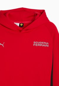Červená mikina s kapucí vyrobená z měkké látky, která obsahuje klokaní kapsu, šedé logo "SCUDERIA FERRARI", kapuci a kontrastní černé boční panely.
