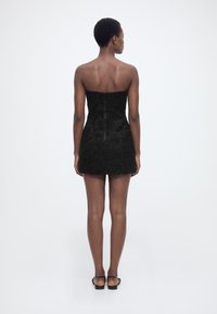 Γυναίκα που στέκεται με την πλάτη, φοράει μαύρο μίνι φόρεμα χωρίς τιράντες με υφή από λουλούδια και μαύρα σανδάλια με λουράκι στον αστράγαλο, μπροστά σε απλό φόντο.