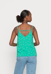 Top de alças finas verde com padrão floral, apresentando um design com costas em V e pequenas flores brancas sobre um fundo texturizado. Combinado com jeans azuis.
