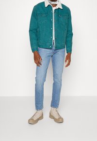 Teal denimjacka med fårskinnskrage, två bröstfickor, knäppning med knappar, kombinerad med ljusblå jeans och beige högstövlar.