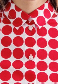 Czerwona i biała koszula w groszki z kołnierzykiem i wyraźnymi guzikami, wykonana z lekkiego materiału o gładkiej teksturze.