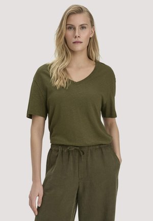 Frau mit langen blonden Haaren, die ein lässiges olivgrünes, kurzärmeliges V-Ausschnitt-Shirt und passende Hosen mit Kordelzug trägt, steht mit einer Hand in der Tasche.