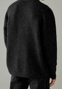 Maglione nero, texturizzato, con una vestibilità ampia, scollatura rotonda e orlo a coste; indossato sopra jeans scuri, mostrando una superficie morbida e pelosa.
