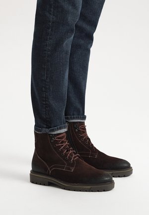 Brązowe zamszowe botki za kostkę z czarną gumową podeszwą, sznurowane, z akcentami stębnowania, noszone z ciemnoniebieskimi dżinsami podwiniętymi na końcach.