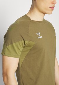 T-shirt à manches courtes vert olive avec un design ajusté, présentant un panneau plus clair sur l'épaule et un logo blanc sur la poitrine.