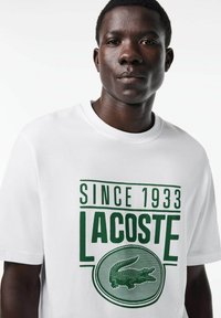 Camiseta blanca de algodón con texto verde "DESDE 1933" y "LACOSTE", con un logo redondo de cocodrilo verde. Mangas cortas, cuello redondo.
