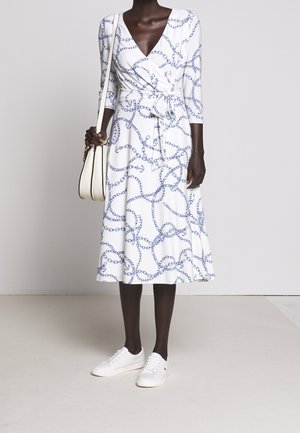 Žena v bílých zavinovacích šatech se vzorem modrých řetězů a kotvicí, v bílých teniskách a s bílou kabelkou přes rameno.