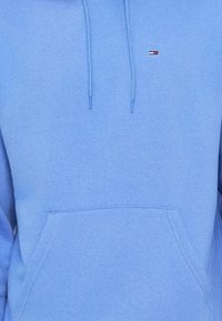 Svetlomodrá mikina s kapucňou vyrobená z jemného materiálu, s prednou kapsou, sťahovacou kapucňou a malým červeným, bielym a modrým logom na hrudi.