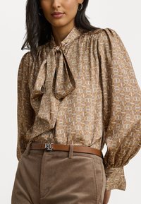 Blusa marrone a fantasia con scollo a fiocco, caratterizzata da un tessuto testurizzato e maniche a sbuffo; indossata con pantaloni di velluto a coste marroni chiari e una cintura.