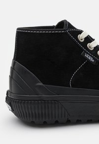 Svart semsket høyhælte sneakers med hvite snører, kontrasterende sømmer, teksturert gummisåle og Vans-logo på siden.