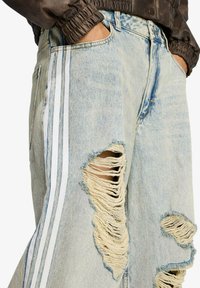 Jeans in denim azzurro chiaro consumato con grandi strappi, strisce laterali bianche e vestibilità comoda. Sfilacciature visibili sui bordi aggiungono texture.