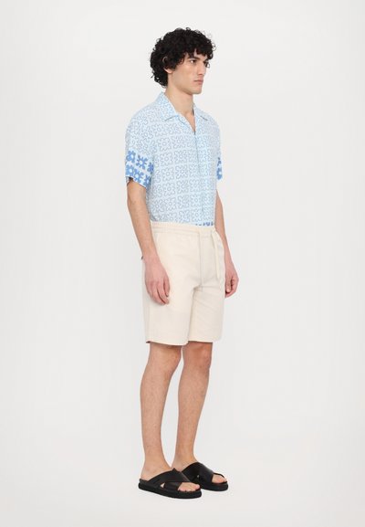 Cămașă cu mânecă scurtă, model floral albastru, pantaloni șort bej și sandale negre tip slide. Material neted, croială lejeră și pantaloni cu talie elastică.