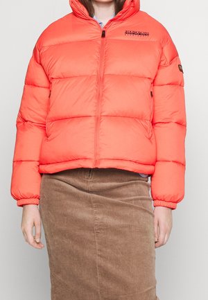 Personne portant une doudoune orange vif avec des poches zippées et une jupe en velours côtelé marron, debout devant un fond uni.