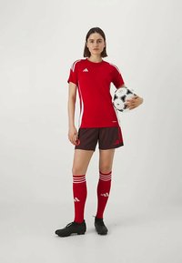 Rød Adidas fotballsjakke med hvite detaljer, sorte shorts, røde sokker med hvite striper, og svarte fotballsko. Holder en svart og hvit fotball.