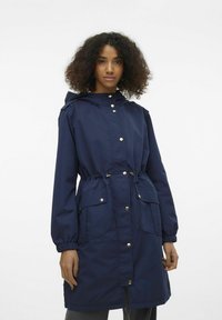 Marineblauwe waterdichte jas met aansnoerbare taille, knoopsluitingen en voorzakken. Beschikt over een capuchon en elastische manchetten voor een strakke uitstraling.