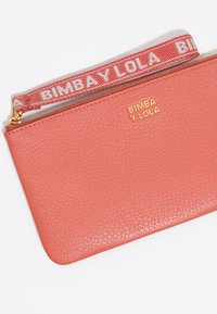 Clutch de cuero coral con un acabado texturizado, logo dorado y una correa de tela de patrón rojo etiquetada "BIMBA Y LOLA". Cierre de cremallera.