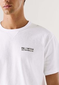 Wit katoenen T-shirt met korte mouwen, voorzien van een ronde hals en zwarte gedrukte tekst aan de linkerkant. De stof voelt zacht en licht gestructureerd aan.