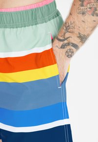 Värikätk swim shortsit, joissa on monivärinen raidallinen kuvio vihreässä, oranssissa, keltaisessa, sinisessä ja tummansinisessä. Varustettu joustavalla vyötäröllä ja sivutaskulla.