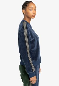 Felpa in velluto navy con strisce laterali testurizzate; presenta un colletto e polsini a coste e un design dal taglio rilassato. Abbinata a pantaloni verdi.