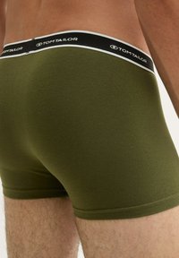 Close-up van een persoon die olijfgroene boxershorts draagt met een zwarte tailleband waarop de merknaam en het logo van Tom Tailor staan.