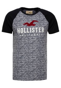 Lyhythihainen t-paita, jossa on harmaaraidallinen vartalo, mustat raglan-hihat ja punainen lintulogoteksti "HOLLISTER CALIFORNIA" valkoisella.