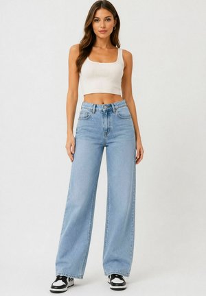 Donna con lunghi capelli castani che indossa un top corto bianco, jeans a gamba larga azzurri a vita alta e sneakers nere e bianche, in piedi.