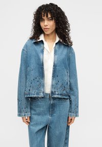 Τζιν μπουφάν με κομψό κοντό σχέδιο, ανοιχτό μπλε χρώμα, διακοσμημένο με μαύρα καρφιά και κολάρο με κουμπιά. Διαθέτει δύο μπροστινές τσέπες.