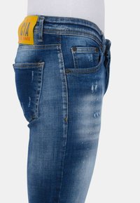 Blauwe vervaagde denim spijkerbroek met lichte slijtage, details op de achterzak en een gele leren label met zwart en rode tekst op de tailleband.