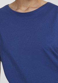 Close-up van een persoon die een eenvoudig blauw crewneck shirt draagt, met zicht op de bovenborst en de halslijn.