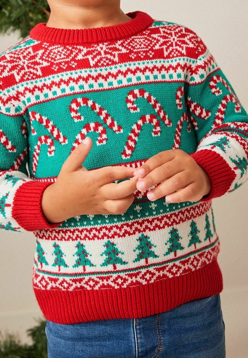 Enfant portant un pull de Noël rouge, vert et blanc avec des motifs de cannes en sucre d'orge et d'arbres de Noël, mains jointes au niveau du ventre.