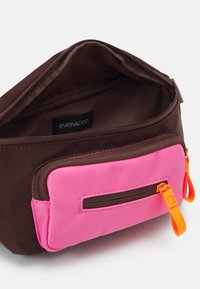 Borsa di stoffa marrone con una tasca frontale rosa, chiusura con zip e dettagli arancioni. L'interno ha una semplice foderatura marrone con un'etichetta del marchio.