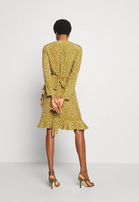 Kvinna med kort krulligt hår som bär en senapsgul blommig klänning knuten i midjan, står med händerna sammanflätade bakom ryggen och har på sig klackskor.