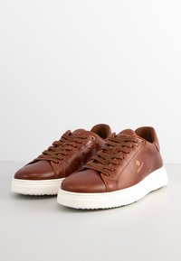 Braune Leder-Sneaker mit einer weißen Gummisohle. Zu den Merkmalen gehören eine abgerundete Spitze, ein Schnürdesign und ein goldenes Logo auf der Seite.