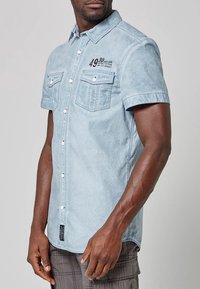 Mies, joka pitää yllään vaaleansinsistä lyhythihaista nappipaitaa, jossa on rintataskut ja painettua tekstiä vasemmalla ylhäällä.