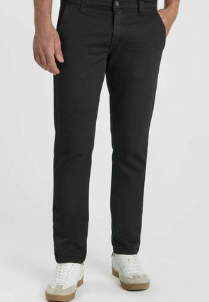 Man die zwarte slim-fit broek draagt met handen in de zakken en witte sneakers op een effen lichte achtergrond.
