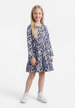 Smilende jente står, iført en langermet mørkeblå kjole med hvitt blomsterprint og hvite høye joggesko på ensfarget bakgrunn.