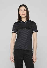 Maillot de sport noir avec un motif en chevron, manches courtes, col en V avec des accents blancs, et un logo imprimé sur la poitrine.