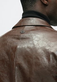 Jachetă maro din piele cu finisaj texturat, pliuri vizibile și un mic logo metalic pe spate. Pulover negru cu guler înalt dedesubt.