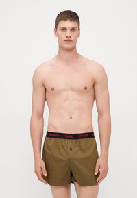 Olivgrüne Boxershorts mit einem elastischen Bund in Schwarz und Rot, der mit "HUGO" beschriftet ist. Leichter Stoff mit einer Knopfverschluss vorne.