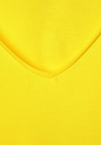 Žlutý top s výstřihem do V vyrobený z měkké, elastické tkaniny. Textura je hladká s jednobarevným provedením, které má čistý, jednoduchý design a žádné ozdoby.
